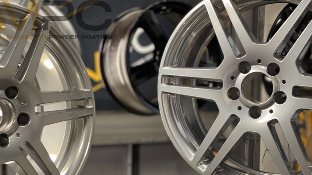 A handful of wheels refurbished in "brilliant silver". They are hanging from the ceiling in a powder coating booth with a sign for manchester powder coating behind them.