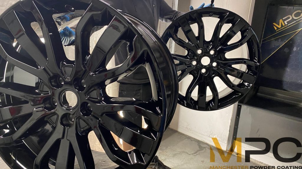 Two wheels refurbished in "gloss black". They are hanging from the ceiling in a powder coating booth with a sign for manchester powder coating behind them.