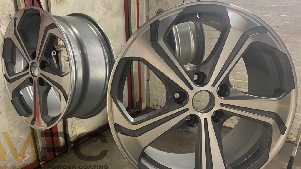 two wheels hanging up in a powder coating booth. They are a light grey colour, with a shiny alloy facing. They are hung up, ready to have a clear lacquer over the top. The wheels are diamond-cut.